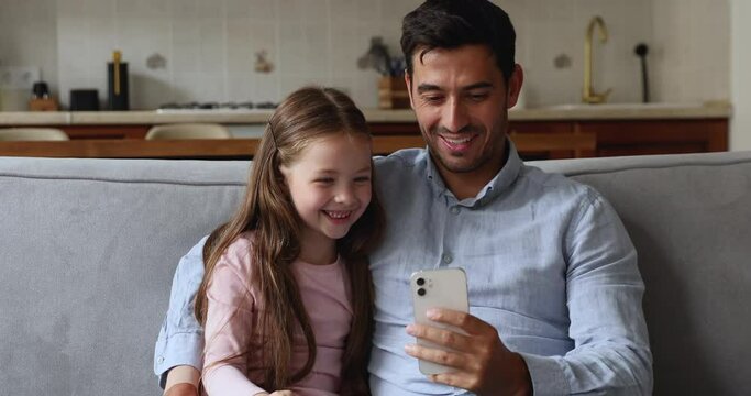 Cheerful Cute Little Daughter Girl And Happy Handsome Dad Speaking On Mobile Phone, Making Video Call, Talking, Laughing In Chat, Conversation, Watching Media Content On Smartphone, Hugging On Couch