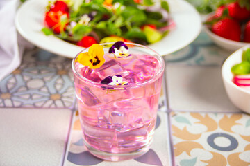 Glasses of pink violet purple cocktail decorated flowers viola pansy and fresh salad on the background.