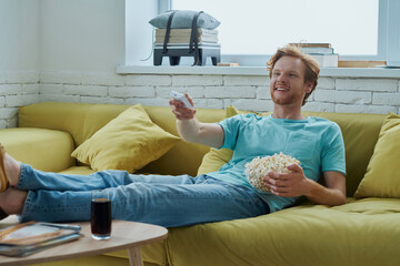 Happy young man watching TV and enjoying snacks while sitting on the couch at home