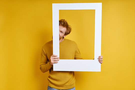 Playful man hiding half face behind a picture frame while standing against yellow background