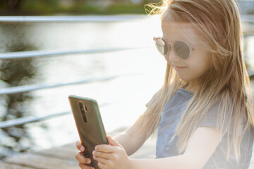 A child talks on a video link on a phone in sunglasses at sunset on the riverbank in the city
