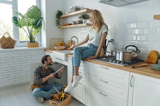 Confident young man repairing a sink while woman sitting on the kitchen counter and smiling