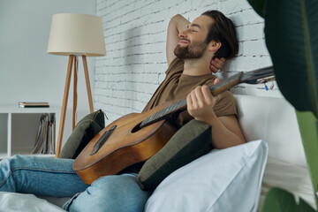 Handsome young man holding acoustic guitar while relaxing on bed at home