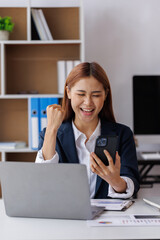 Happy Excited Asian young entrepreneur business woman using phone and laptop sitting on a desk officer in the day at the office