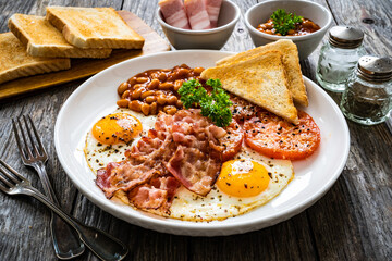 English breakfast - toasts, sunny side up eggs, bacon ,beans in sauce and fried tomato on wooden table
