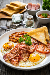 English breakfast - toasts, sunny side up eggs, bacon ,beans in sauce and fried tomato on wooden table
