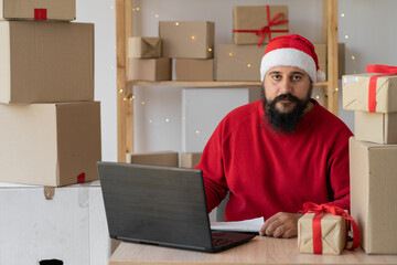 small business owner Indian freelance Santa working in home office using computer, online marketing packaging box and gifts for Christmas delivery, SME e-commerce