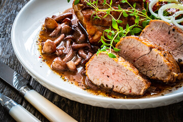Roasted pork loin with shimeji mushrooms in sauce, fresh thyme, greens and cherry tomatoes on wooden table
