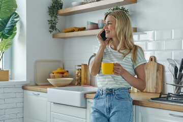 Attractive young woman talking on mobile phone while enjoying coffee at the kitchen