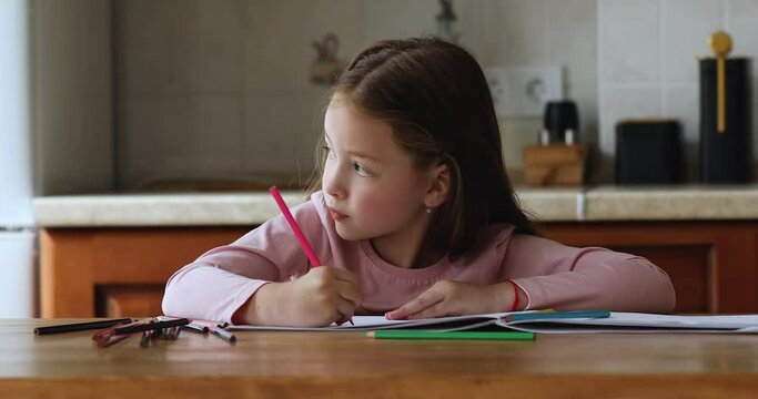 Focused sweet primary school kid doing homework, learning to write, drawing doodles in paper album, sitting at kitchen table with colorful pencils, looking at window, turning head away, smiling