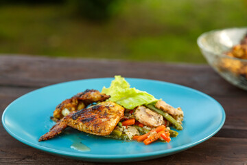 A plate of chicken with vegetables on a wooden table next to a dish with grilled wings.