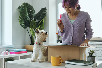 Beautiful young woman unpacking box while her cute dog sitting near her on the table