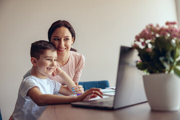 Small cute schoolboy with braces using laptop in online learning, happy mother smiling.