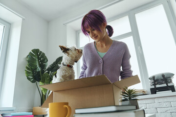 Beautiful young woman unpacking box while her cute dog sitting near her on the table