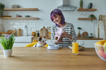 Cheerful young woman feeding her little dog while sitting at the kitchen island