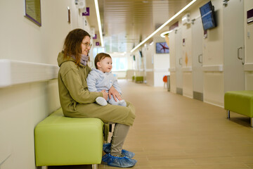 Mother woman with toddler baby boy in her arms visit in the hall clinic. Mom with a child in the corridor of the hospital waiting for a doctor appointment. One year old kid