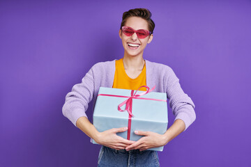 Excited young woman carrying gift box while standing against purple background
