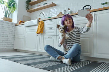 Young woman carrying little dog and making selfie while sitting on the floor at the kitchen