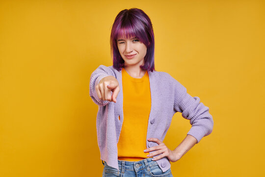 Beautiful young woman pointing camera and winking while standing against yellow background