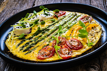 Tasty breakfast. Omelette  with green asparagus, goat cheese, pepper and cherry tomatoes served on black plate on wooden table
