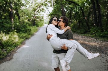 A bearded long-haired stylish man holds his beloved hippie brunette woman in sunglasses in his arms and circles her in nature in the park. Portrait, photo of happy and smiling newlyweds.