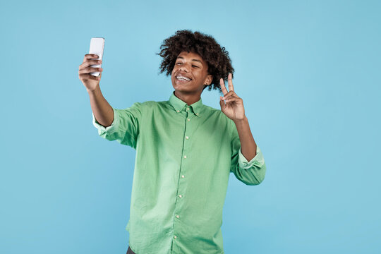 Excited african american guy taking selfie on smartphone and showing v-sign, streaming live on web, blue background