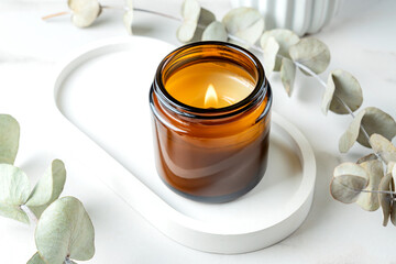 Burning candle in amber glass jar on concrete trendy tray and eucalyptus branches on white marble...