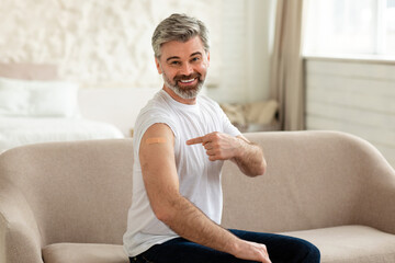 Happy Vaccinated Man Showing Arm With Plaster Sitting At Home