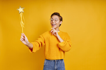 Joyful young woman holding magic wand while standing against yellow background