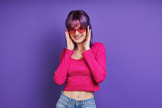 Cheerful young woman adjusting eyeglasses while standing against purple background