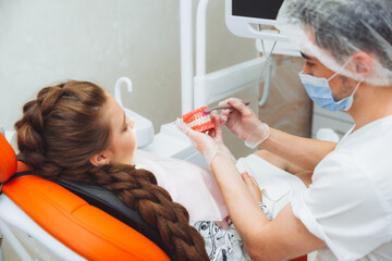 a male dentist in a medical uniform shows a little girl how to properly brush her teeth on a jaw...
