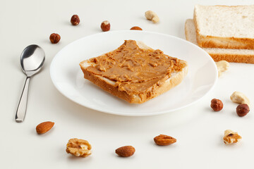 Square bread for toast with peanut butter on a plate. Nuts, a spoon, slices of bread and a white plate with a sandwich on a white table.