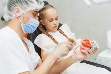 a male dentist in a medical uniform shows a little girl how to properly brush her teeth on a jaw...