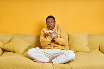 Terrified man holding plate with popcorn while sitting on the couch with yellow background