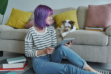 Cheerful teenage girl using digital tablet while her little dog sitting near her on the couch