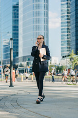 A female realtor in a blazer is walking with a laptop and having a conversation on a cell phone between the office buildings in the financial district. A businesswoman in a cluster of tall buildings.