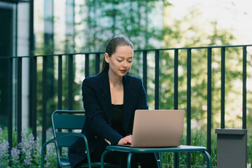 A female realtor in a blazer is working on a laptop in a modern urban space in the financial district. A businesswoman is sitting at the table in the contemporary garden in a cluster of tall buildings