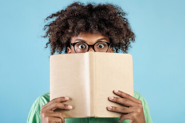 Portrait of emotional african american guy in glasses covering face with book, hiding behind it...