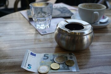 Tisch mit Glas, Tasse, silbernem Aschenbecher, Geldschein und Euro-Münzen in Café in Stadt 