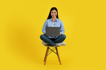 Cheerful young chinese lady student sitting on chair and working on laptop, isolated on yellow background