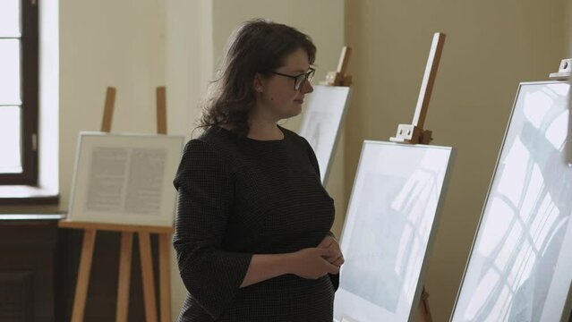 Young plus size, overweight thoughtful woman with freckles in glasses carefully examines unrecognizable paintings in art gallery at an exhibition. Reflection of the window in the drawings of artists.