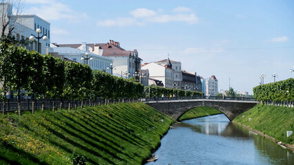 Beautiful cityscape with a narrow canal and two slopes covered by green grass on blue cloudy sky background. Stock footage. Summer city street with white houses, and people walking on the bridge.