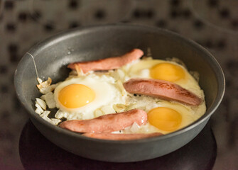 fried fried eggs with sausages and onions in a frying pan on an electric stove.
