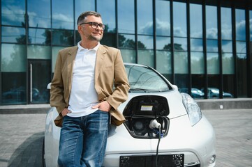 Casual man near electric car waiting for the finish of the battery charging process.