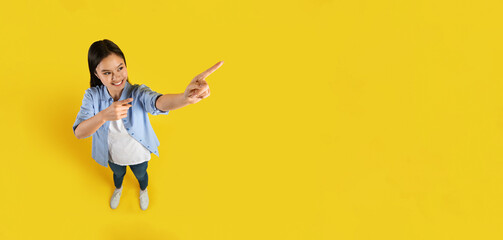 Glad happy positive young asian female student show fingers up to empty space, isolated on yellow background