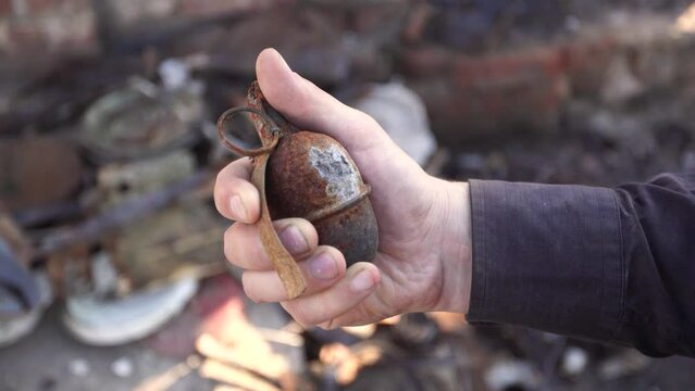 Close up view 4k stock video footage of adult male hand holding real burnt out unexploded hand grenade isolated on blurry debris and ruins of civil house after been bombed by russian aircraft. Ukraine