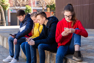 Teen male and female students sitting together outdoor ignoring each other while browsing internet on smartphones - obrazy, fototapety, plakaty