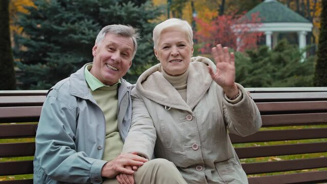 Caucasian family couple lovely elderly married grandparents sit on bench in park wave hands welcome sign hello goodbye gesture greeting looking at camera senior man husband hugging mature woman wife