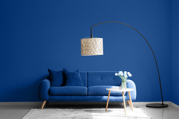 Interior of living room with comfortable sofa, floor lamp and table with flowers near blue wall
