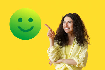 Beautiful young woman pointing at smiling emoticon on yellow background